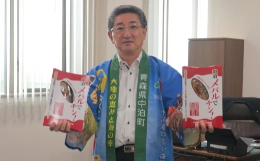 中泊町の青い法被を着た濱舘町長が町の特産品「中泊メバルでチン!」を両手に持っている写真