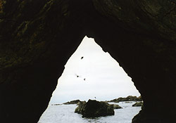 暗い洞窟の中から見える三角に切り立った入口の奥に、小島がある海が見えている写真