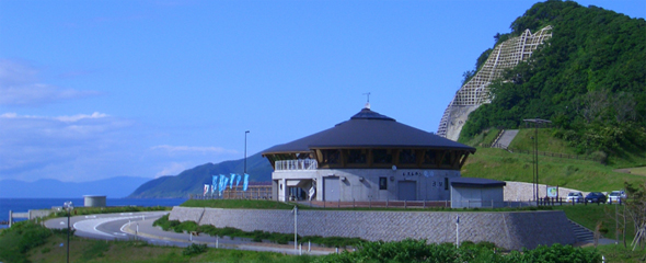 快晴の青空が広がる中、海と山を背景にして中央に建つ道の駅全景の写真