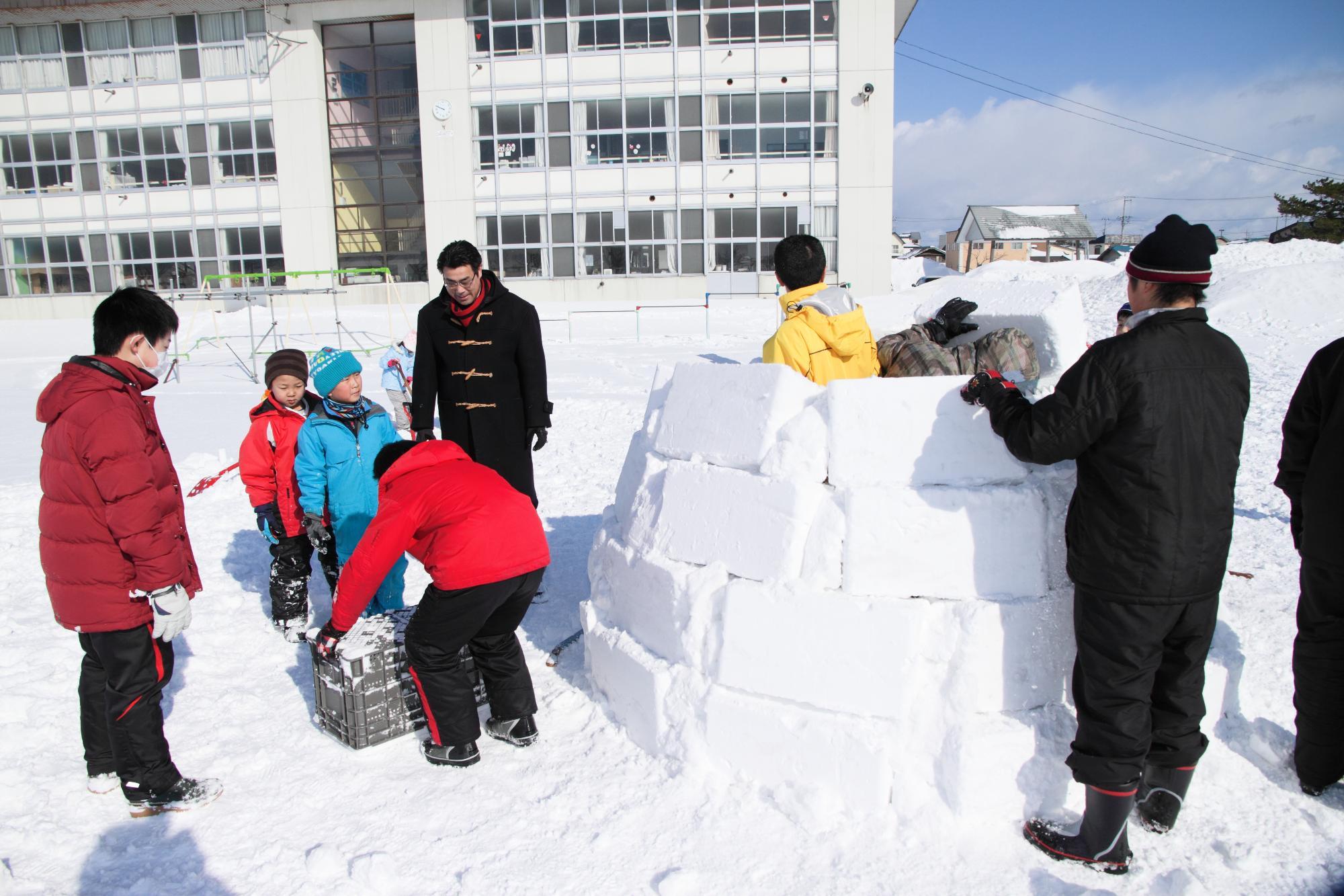 雪が積もった学校の校庭で、大人2人と子ども6人が四角い雪の塊を積み上げてイグルーを作っている写真