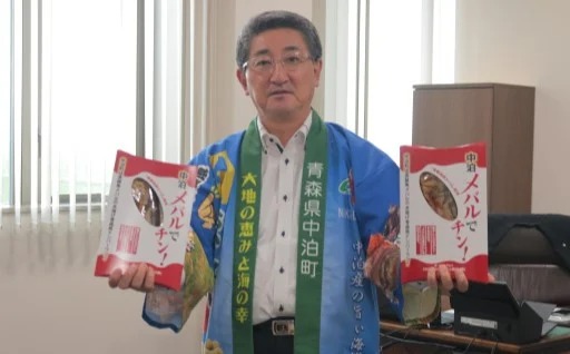 中泊町の青い法被を着た濱舘町長が町の特産品「中泊メバルでチン！」を両手に持っている写真